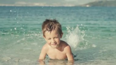 Denizde yüzen ve gülümseyen bir çocuğun yavaş çekim videosu. Yaz tatilindeki çocuk sahildeki okyanusta mutlu bir şekilde yüzüyor..