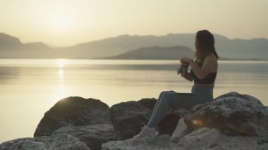 Kız kahve içiyor. Deniz kenarında koştuktan sonra termal bardaklar. Gün batımı ve spordan sonra dinlenen güzel bir kadın. Yüksek kalite 4k görüntü