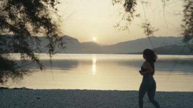 Caucasian girl on a morning jog by the ocean. Girl at sunrise runs on the beach near the waves of the sea with a background of silhouetted mountains.