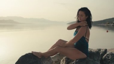 Beautiful Caucasian girl at sunrise sitting on a rock by the ocean. Dreamy woman looks at the sunset on vacation by the sea, lifestyle to relax.