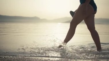 Gün doğumunda sahilde yürüyen ve ayaklarını okyanus suyunda ıslatan bir kızın yavaş çekim videosu. Denizin kenarında yürüyen dişi bacakların yakın çekimi. Yüksek kalite 4k görüntü