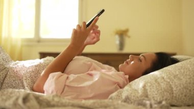 The girl turns off the alarm clock in the morning in bed and gets up. A beautiful woman in a shirt is texting on a gadget in the morning rays of the sun. High quality 4k footage