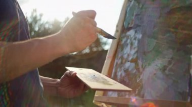 A male artist paints a picture on an easel with a brush. Beautiful sunset in nature, inspiration tree art and hobby drawing. High quality 4k footage