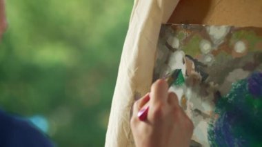 A close-up of a brush in the hands of a l man, with which he paints a picture. Relaxation in nature, painting on an easel with paints. High quality 4k footage