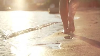 Close-up footage of female legs walking on the sandy beach at sunset near the sea. Ocean waves, sunlight, relaxation and recreation. High quality 4k footage