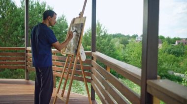 A male artist stands on the terrace at home and paints a picture with a brush. The art and hobby of easel painting. High quality 4k footage