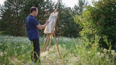 The artist paints a picture against the background of nature. A man is looking for inspiration with a brush and an easel in a beautiful landscape of flowers. High quality 4k footage