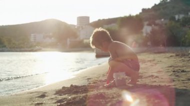 A boy plays with a bucket and makes figures from sand on the beach by the sea. A child plays on the ocean shore with sand and toys. High quality 4k footage