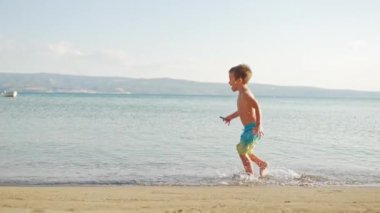 Happy and smiling boy running on the beach near the sea. The child is happy to rest on the ocean shore, running on the water. High quality 4k footage