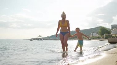 A mother and son walk holding hands on the seashore, splashing water with their feet. Happy family smiling together on the ocean shore, slow motion video. High quality 4k footage