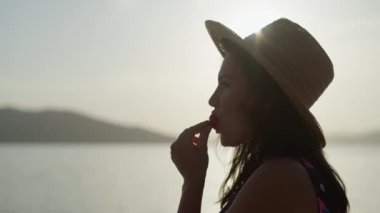 Bir kız gün batımında deniz kenarında bir çileği ısırıyor. Güzel bir kadın yazın okyanusun kenarında dinlenir ve meyve yer. Yüksek kalite 4k görüntü