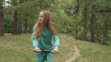 Gülümseyen bir kız parkta scooter sürüyor. Doğada aktif eğlence, raylarda gitmek. Mutlu çocuk. Yüksek kalite 4k görüntü