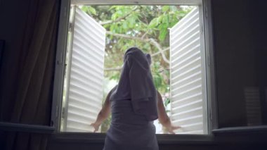 Havlulu güzel bir genç kız pencereyi açıyor. Evde güneşli güzel bir sabah. Yeni bir günün başlangıcı ve romantik bir kadın. Yüksek kalite 4k görüntü