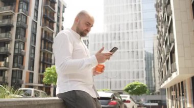 Kahve molasında olan bir iş adamı iş merkezinin yanındaki telefonda mesajlaşıyor. Bir adam olumlu duygularla gülümser. Yüksek kalite 4k görüntü