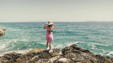 Deniz kenarında gezintiye çıkmış şapkalı bir kız duruyor ve dalgalara bakıyor. Güzel bir kadın sadece okyanusun kenarında dinlenir. Yüksek kalite 4k görüntü