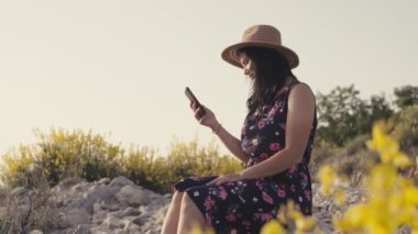 Dağlarda bir taşın üzerinde otururken şapkalı bir kız akıllı telefondan yazıyor. Bir kadın sevgilisine mesaj atıyor ve gülümsüyor. Yüksek kalite 4k görüntü