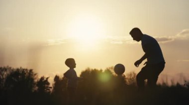 Parkta futbol oynayan bir adamla bir çocuğun yavaş çekim videosu. Baba ve oğlunun siluetleri parkta top oynayarak vakit geçiriyorlar. Mutlu ve sevgi dolu bir aile. Yüksek kalite 4k görüntü