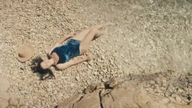 Güzel beyaz kadın deniz kenarında çakıl taşı plajında güneşleniyor. Kız rahatlıyor, denizin dalgaları ayaklarını güzel bir manzarada yıkıyor. Yüksek kalite 4k görüntü