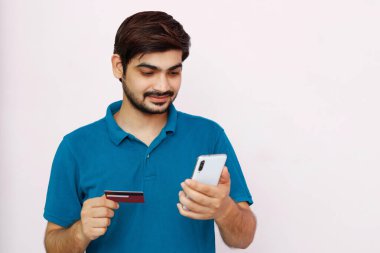 Yakışıklı adam, kredi kartı ve akıllı telefon kullanarak ödeme yapıyor. İzole edilmiş arka planda Hintli tişörtlü adamın fotoğrafı..