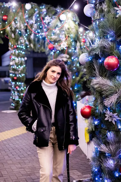 A girl in a coat and gloves stands under a Christmas tree decorated with lights. New Year 2022.