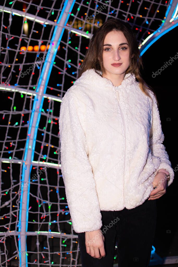 A girl in a coat and gloves stands under a Christmas tree decorated with lights. New Year 2022.