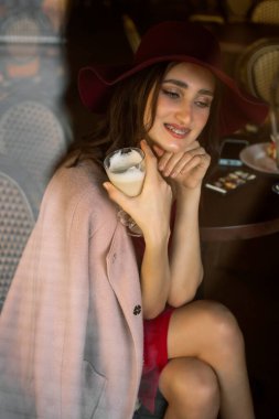 The girl eats a donut and drinks coffee. A young brunette girl in a red dress eats a pink donut in a cafe for breakfast.