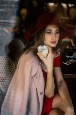 The girl eats a donut and drinks coffee. A young brunette girl in a red dress eats a pink donut in a cafe for breakfast.