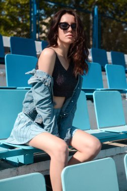 A brunette in a blue bow sits on the bench of football fans. A brunette in a blue skirt and top sits on a blue cheerleader bench.