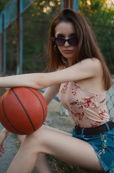 細い女の子がボールを手にバスケットボール場に腰を下ろしている ブルネットーネットとともにAボールで彼女の手 — ストック写真