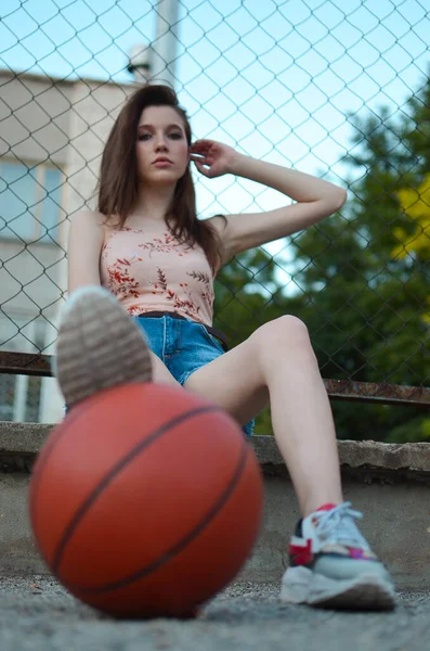 一个身材苗条的女孩手里拿着一个球坐在篮球场上 一个带着雪茄酱的黑发女人坐在那里 手里拿着一个球 — 图库照片