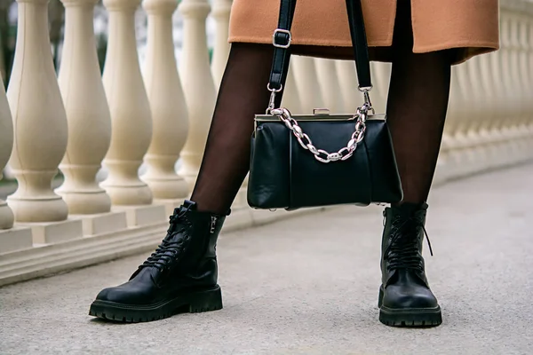 Stylish look. Close-up of a black handbag in the hands. Boots are black. Fashionable girl on the street. Close-up photo of a girl\'s legs and a handbag in her hands.
