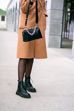 Elinde bir torba var. Elinde çanta tutan bir kız. Bej arka planda siyah bir çanta. Bej ceket. Zincirli siyah çanta. Kızın bacaklarındaki siyah ayakkabıların yakın çekimi. Deri bir takımın arka planında çanta..