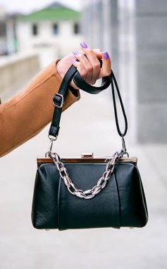 Elinde bir torba var. Elinde çanta tutan bir kız. Bej arka planda siyah bir çanta. Bej ceket. Zincirli siyah çanta. Kızın bacaklarındaki siyah ayakkabıların yakın çekimi. Deri bir takımın arka planında çanta..