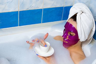 Kafkasyalı genç kızıl saçlı kadın banyoda dinleniyor, kafasının etrafına sarılmış banyo havlusu giyiyor, yüzünde pembe kilden yüz maskesi olan köpük bir küvette uzanıyor, manikürünün tadını çıkarıyor..