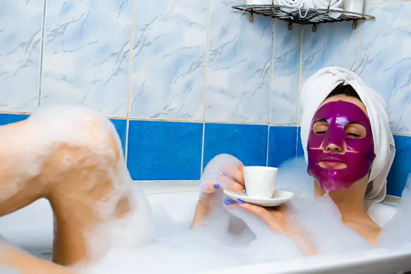 白种人年轻的红头发女人躺在浴室里休息 头戴浴巾 躺在泡沫浴缸里 脸上戴着粉色粘土面罩 享受指甲修剪的乐趣 — 图库照片