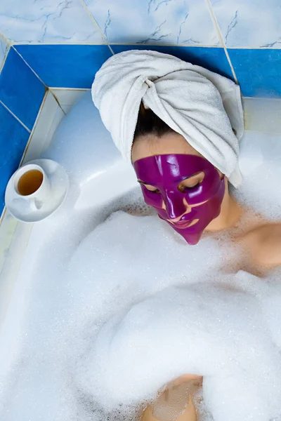 白种人年轻的红头发女人躺在浴室里休息 头戴浴巾 躺在泡沫浴缸里 脸上戴着粉色粘土面罩 享受指甲修剪的乐趣 — 图库照片