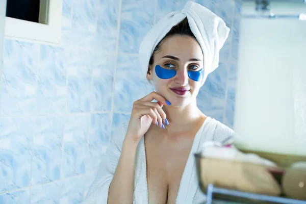 一个穿着浴衣头戴毛巾的女孩在镜子前的浴室里把眼罩贴在了眼皮底下 — 图库照片