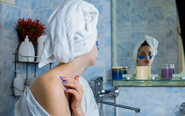 一个穿着浴衣头戴毛巾的女孩在镜子前的浴室里把眼罩贴在了眼皮底下 — 图库照片