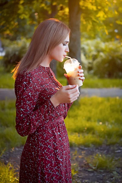 公園を歩いている女性 カップからコーヒーを飲む 背景に美しい夕日 — ストック写真