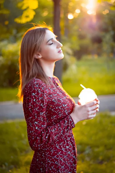 公園を歩いている女性 カップからコーヒーを飲む 背景に美しい夕日 — ストック写真