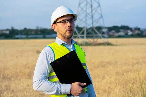 engineer in a helmet, glasses and a yellow vest, hand on a helmet, holding a notebook, a folder, making notes in a notebook a caucasian white engineer