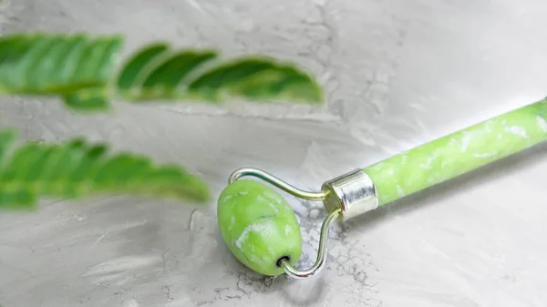 Green jade roller for facial massage and a green branch on a marble background. Accessories for home and personal care. Roller for the face against age wrinkles. Top view, flat lay.