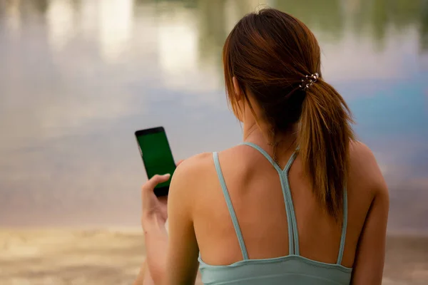 a girl in a sports uniform holds a phone in her hands, dressed in a green tracksuit top and bicycle shorts, water balance, sports activities on the street, sports lifestyle, yoga in the park, doing stretching near the water