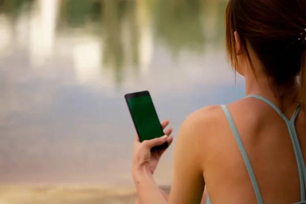 a girl in a sports uniform holds a phone in her hands, dressed in a green tracksuit top and bicycle shorts, water balance, sports activities on the street, sports lifestyle, yoga in the park, doing stretching near the water