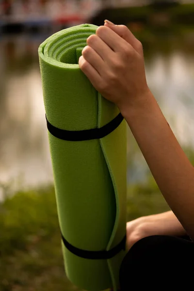 A girl in a sports uniform holds a yoga mat in her hands, dressed in a black sports top, a sports mat in green, yoga in the park, sports lifestyle, outdoor sports