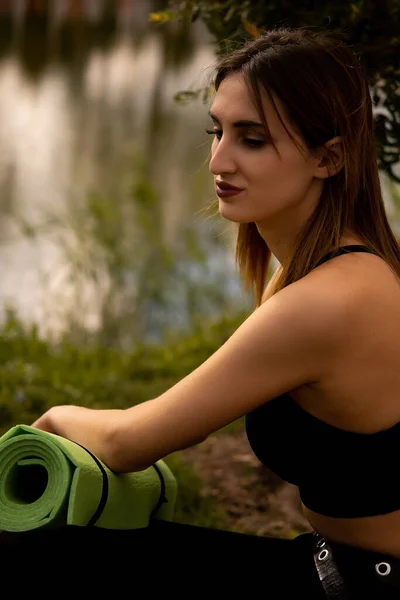 A girl in a sports uniform holds a yoga mat in her hands, dressed in a black sports top, a sports mat in green, yoga in the park, sports lifestyle, outdoor sports