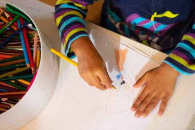 Üst görünüm küçük eller kalem tutar ve beyaz A4 kağıdına kalem dolu yuvarlak bir kutuyla izole edilmiş masa üzerine yüz çizer. Eğitim ve öğrenme süreci kavramı