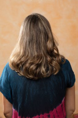 Arkaya bakan esmer, uzun kahverengi düz saçlı, kıvırcık saçlı kadın sarı arka planda duruyor.