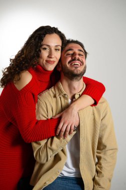 Gri beyaz arka planda izole edilmiş mutlu genç çiftin portresi. Kırmızı elbiseli genç kız, ortağına arkadan sarılıyor.