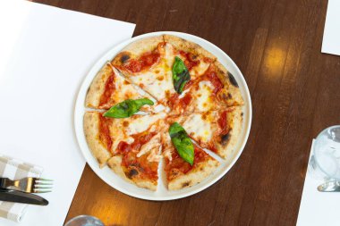 Tahta bir masada beyaz bir tabakta pizza. Çatallı, bıçaklı ve peçeteli leziz peynirli pizzanın üst görüntüsü. İnce hamurlu pizza, İtalyan mutfağı..